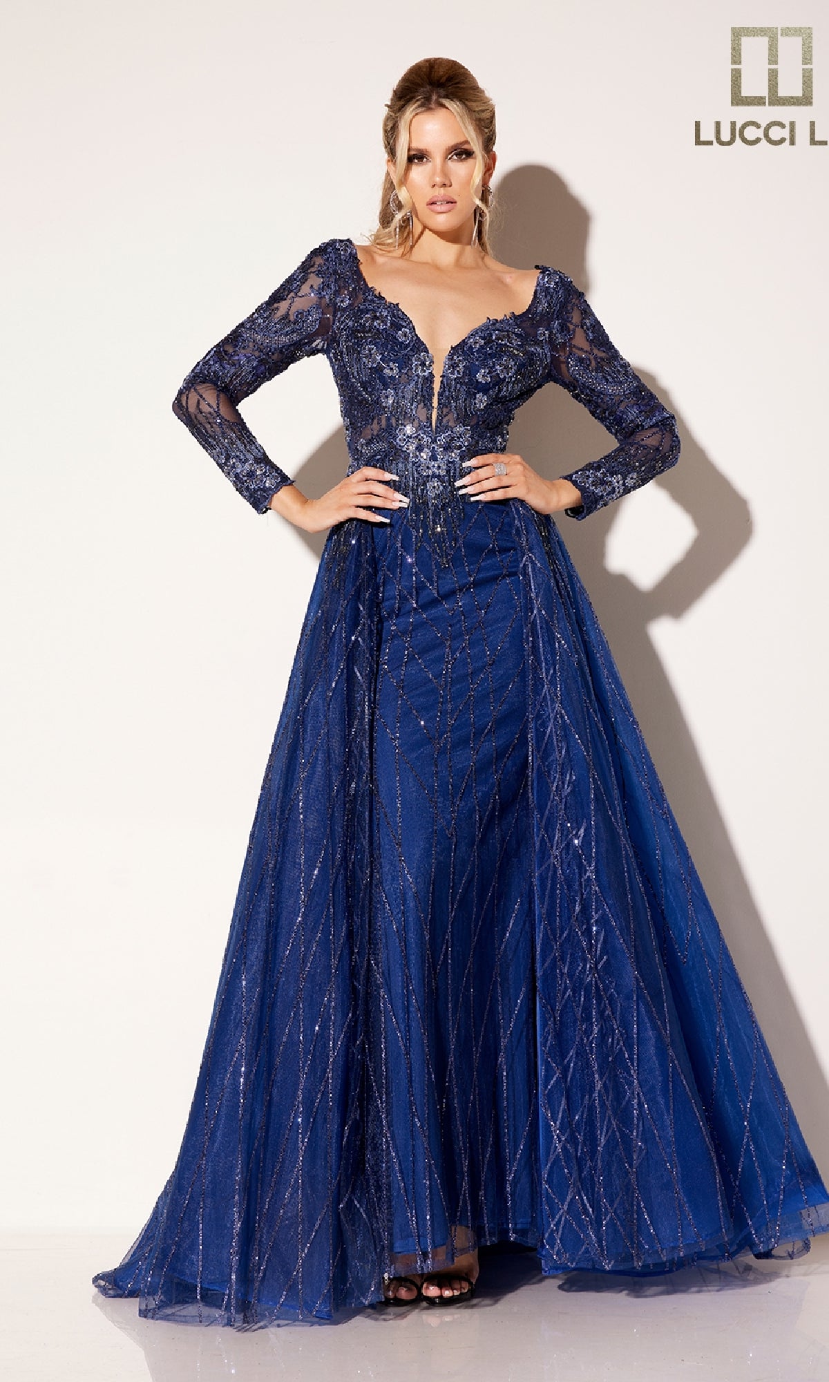 A woman stands against a plain background wearing the Lucci Lu C8076 long prom dress, featuring navy blue sequins, long sheer sleeves, and a plunging v-neckline.